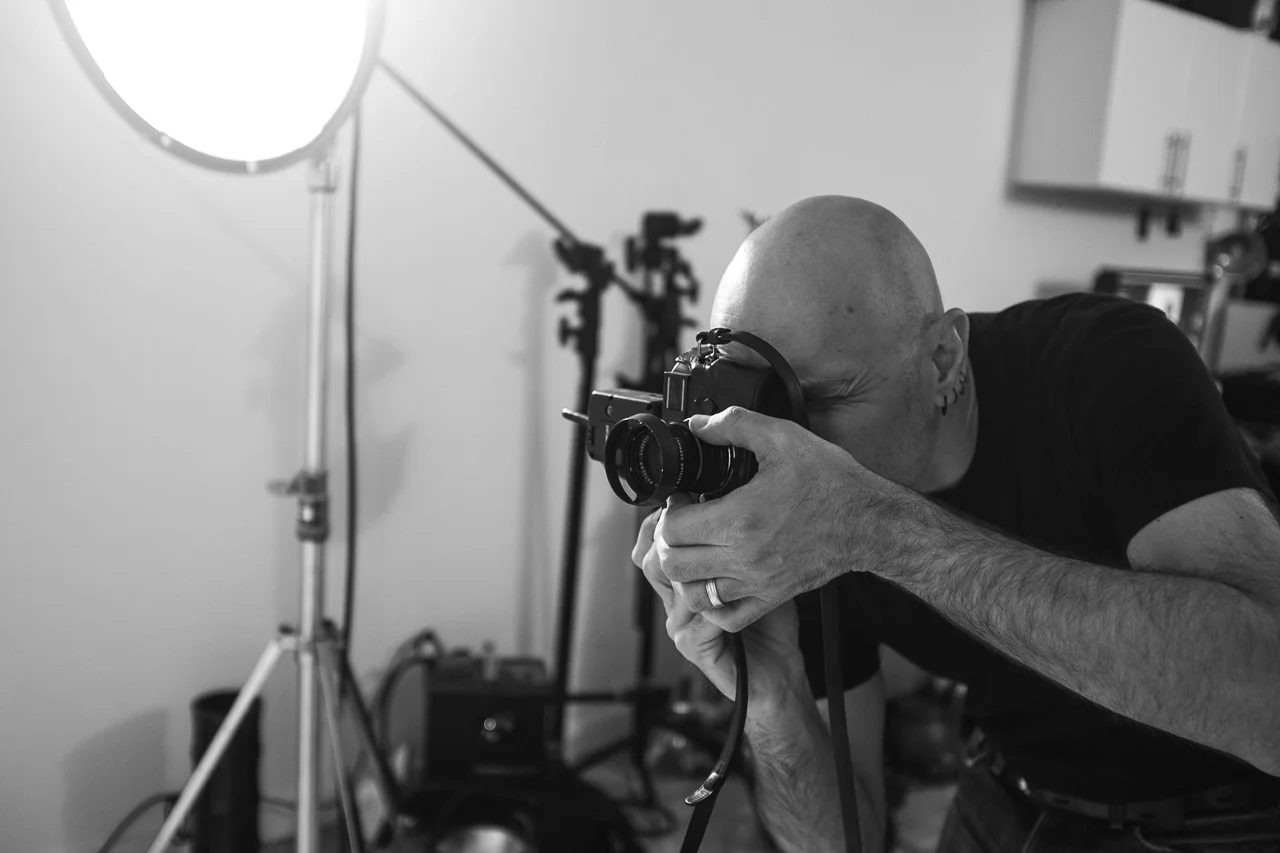 A man with a shaved head and a tattoo on his neck taking a black and white photo indoors, with photography equipment including a large softbox light and tripods in the background.