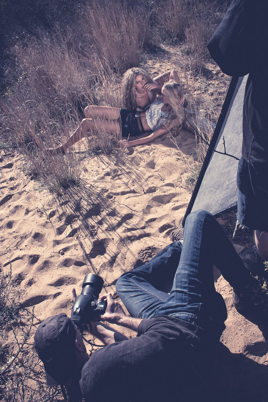 Photoshoot of a woman and a child lying on the sand in a field of tall grass, with a photographer taking their picture and a large reflector.