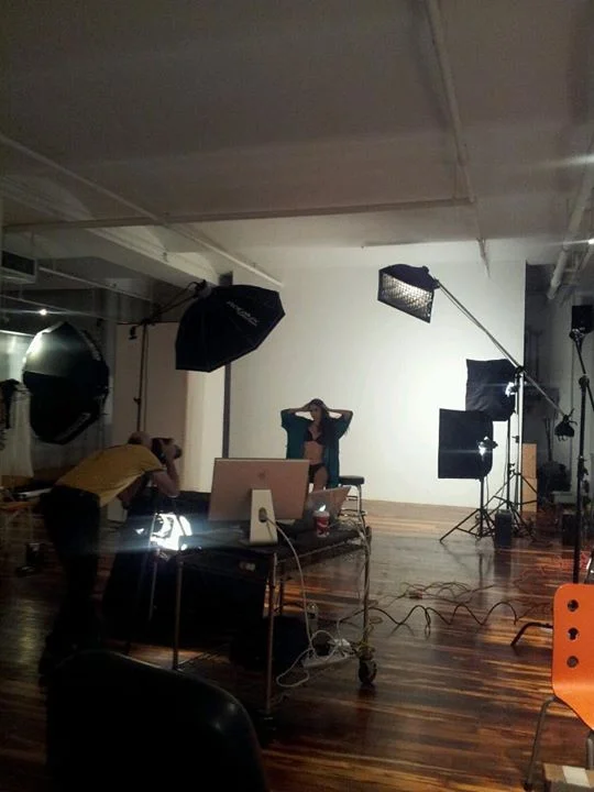 A room with photography studio equipment, including large softboxes, light stands, and a white backdrop wall. Two people are setting up or working in the studio, one adjusting their clothing in front of the backdrop.