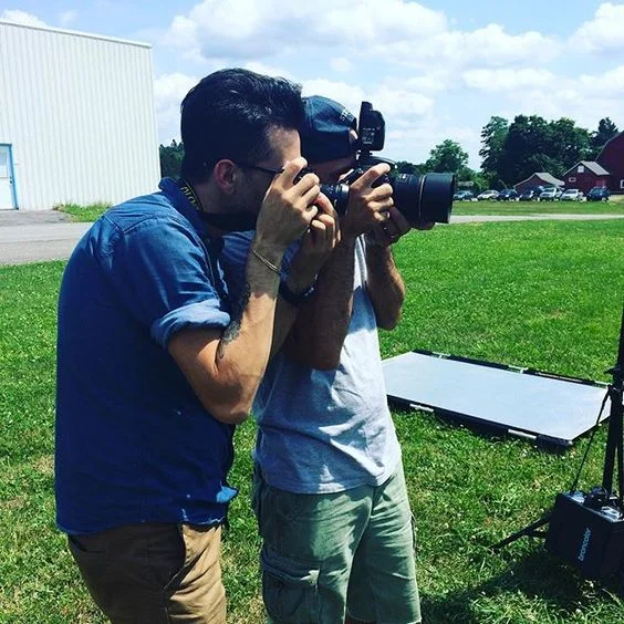 Two men are outdoors, one is taking a photograph with a camera and the other is adjusting a camera on a tripod, on a grassy field with a building and trees in the background.