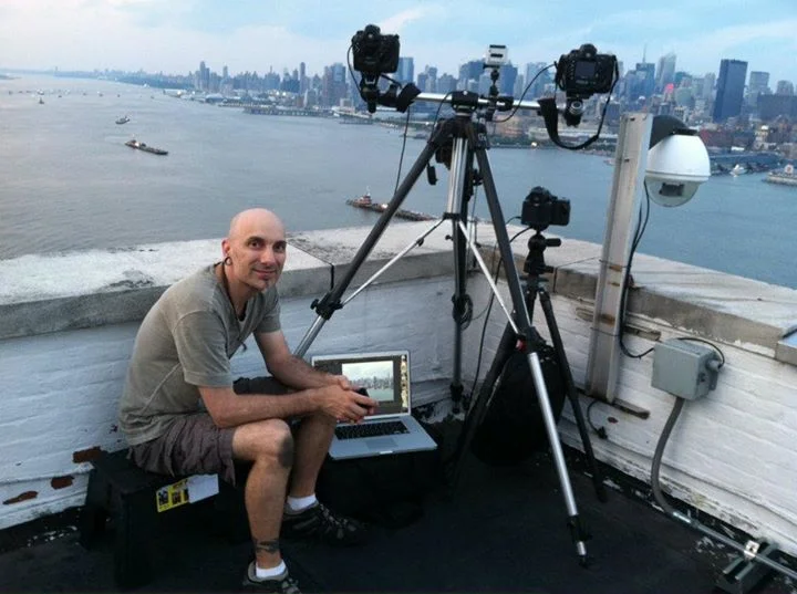A man sitting on a rooftop with skyline and river view, operating a camera setup with two cameras mounted on tripods, a laptop, and other equipment.