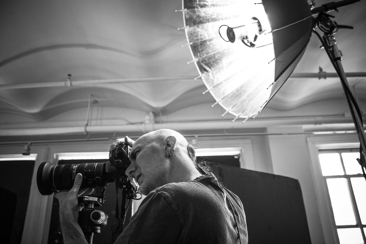 A photographer is looking through a camera on a tripod in a studio with large windows, professional lighting equipment overhead, and a backdrop in the background.