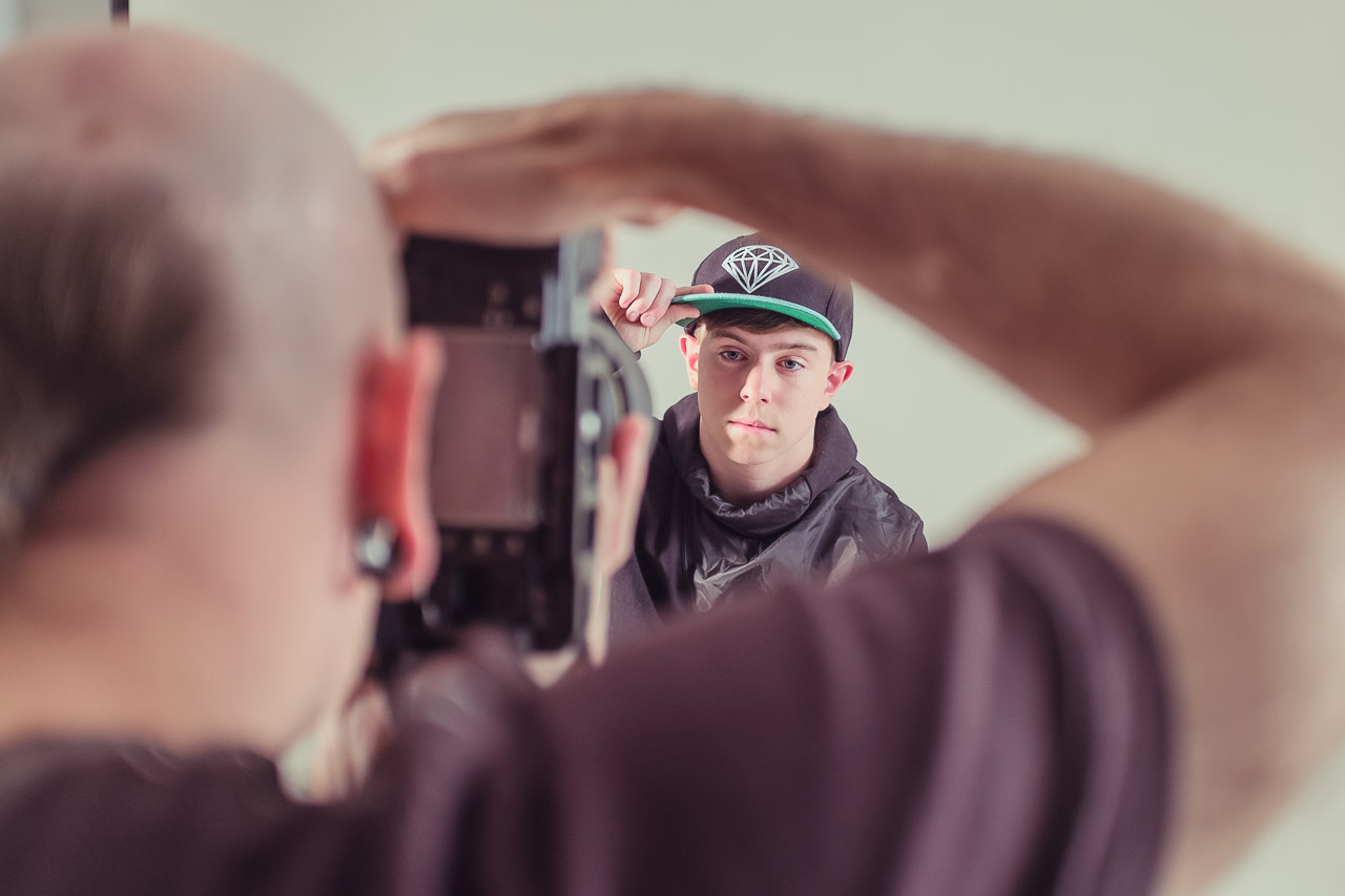 Person taking a photo of a young man wearing a black cap with a diamond logo and a black jacket, posed against a plain background.