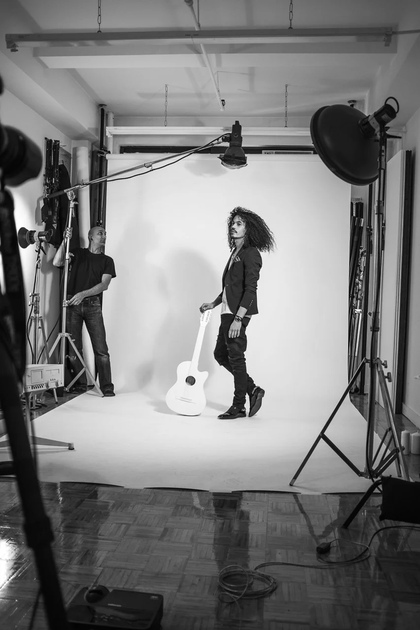 A black and white photo of a photo shoot studio with a woman with curly hair holding a white guitar, standing on a white background, and a photographer or assistant adjusting lighting equipment.