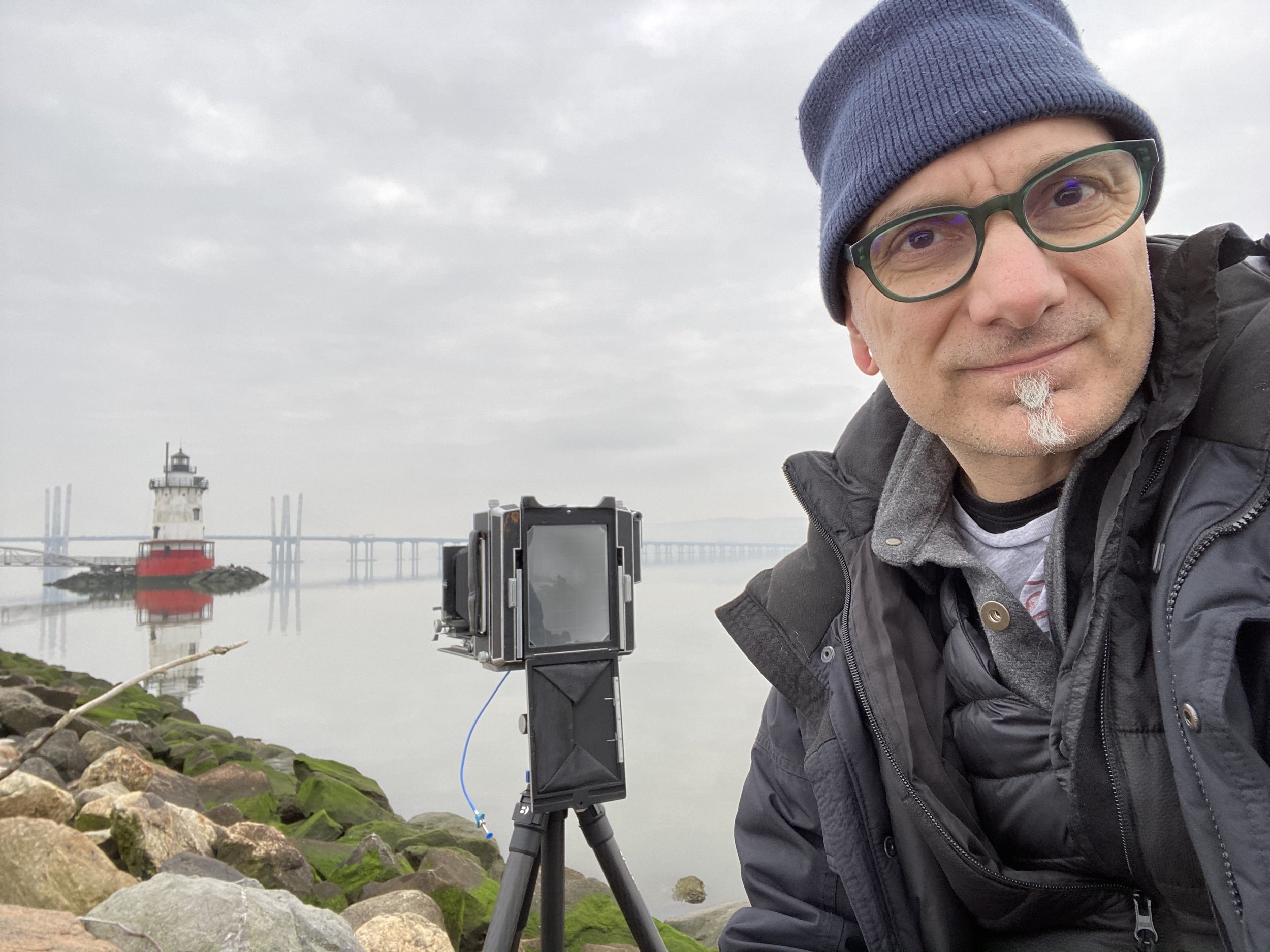 A man taking a selfie by a waterfront with a lighthouse, rocks, and a bridge in the cloudy background. He is wearing glasses, a blue beanie, and a black jacket, with a camera on a tripod in front of him.