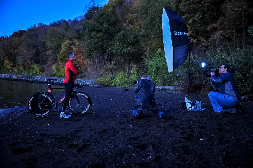 A woman in a red jacket standing with a bicycle on a muddy beach, being photographed by two photographers with professional equipment, near a body of water with a forested hill in the background during dusk.