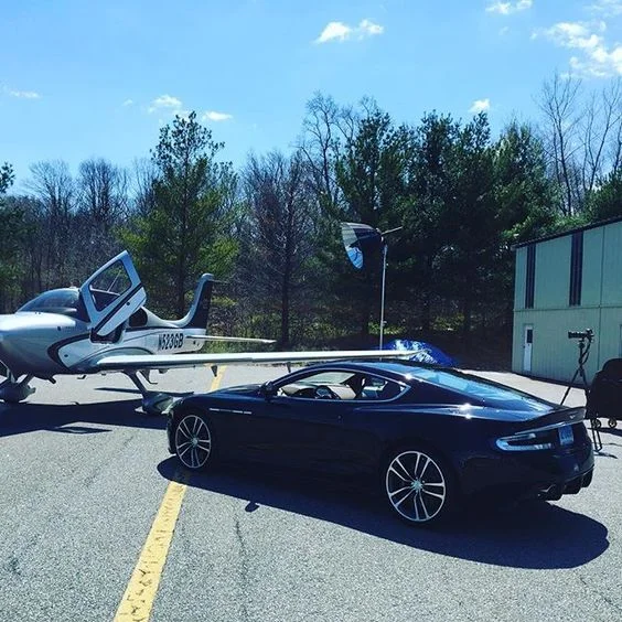 A black sports car parked beside a small private jet plane in an outdoor parking lot, with trees and a building in the background.
