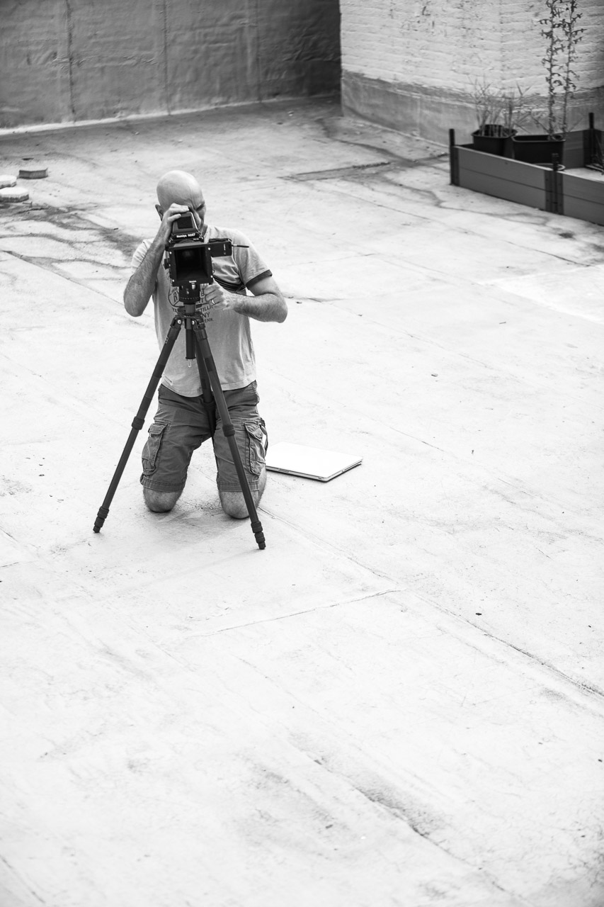 A person kneeling on the ground using a camera mounted on a tripod, with a notebook nearby, outdoors in an urban setting, in black and white.