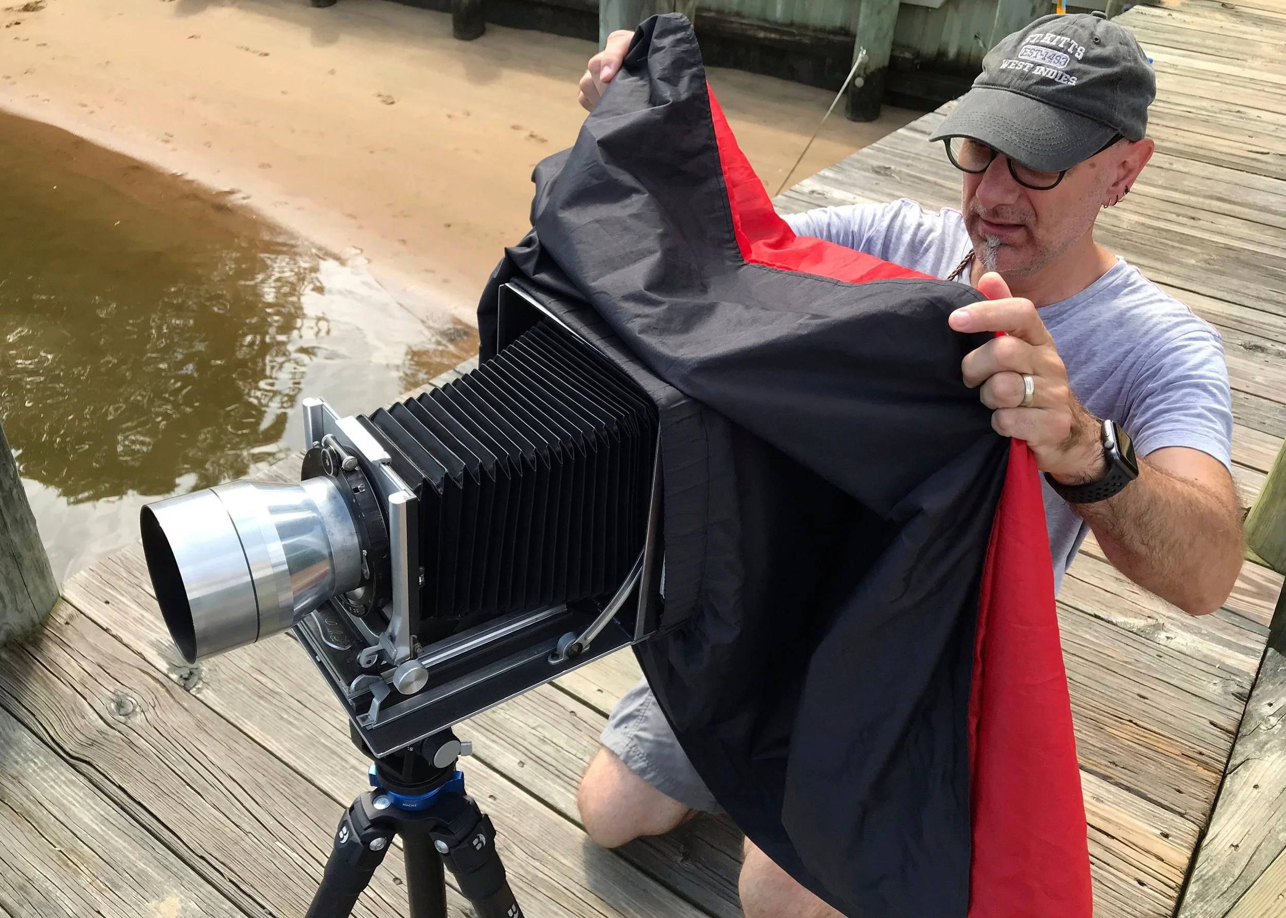 Man setting up large camera on a tripod near a wooden dock by the water, adjusting a black and red camera cover.