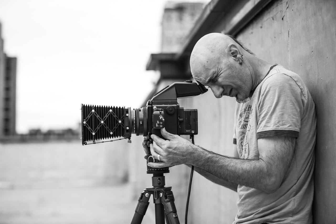 A man with a shaved head and an earring is looking through a camera with a large lens mounted on a tripod, standing against a concrete wall outdoors.