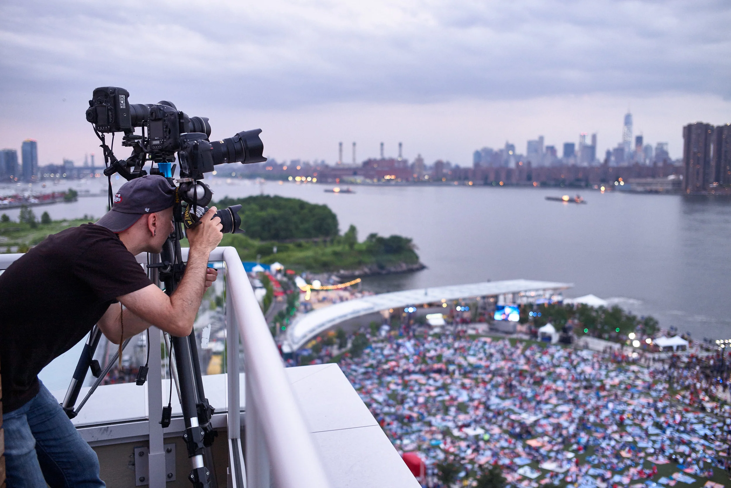 A man operating a camera on a tripod on a rooftop overlooking a large outdoor event near a river with a city skyline in the background during evening.