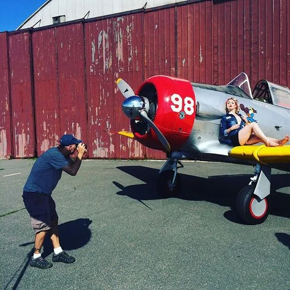 A woman lying on a vintage jet aircraft with the number 98 painted on its nose, posing for a photo as a person takes her picture.