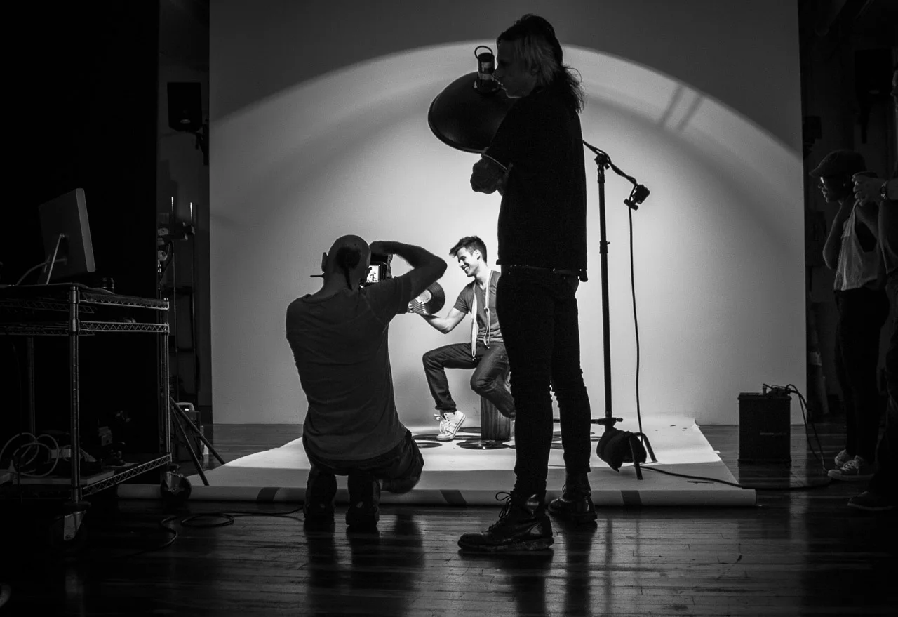 Behind-the-scenes of a photo shoot with a man sitting on a stool, smiling, while a photographer takes a picture and a woman holds a large reflector, with lighting equipment and crew members around.