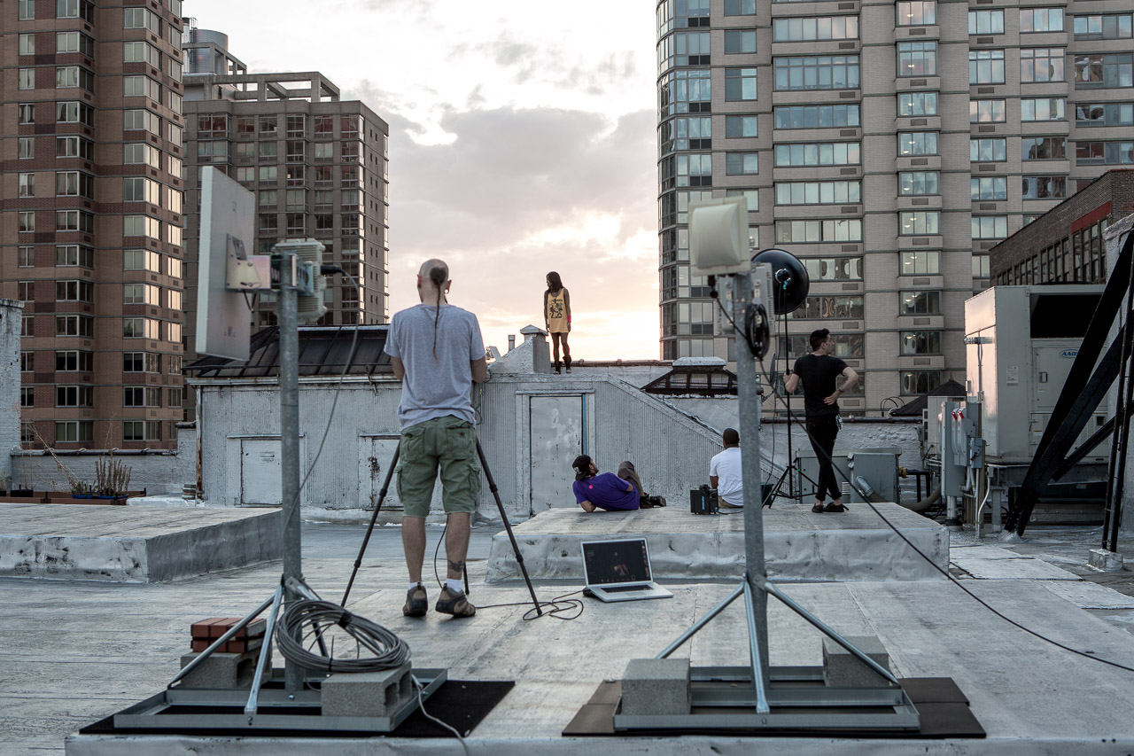 Filmmakers and crew shooting a scene on a city rooftop at sunset, with people sitting and observing, surrounded by tall buildings.