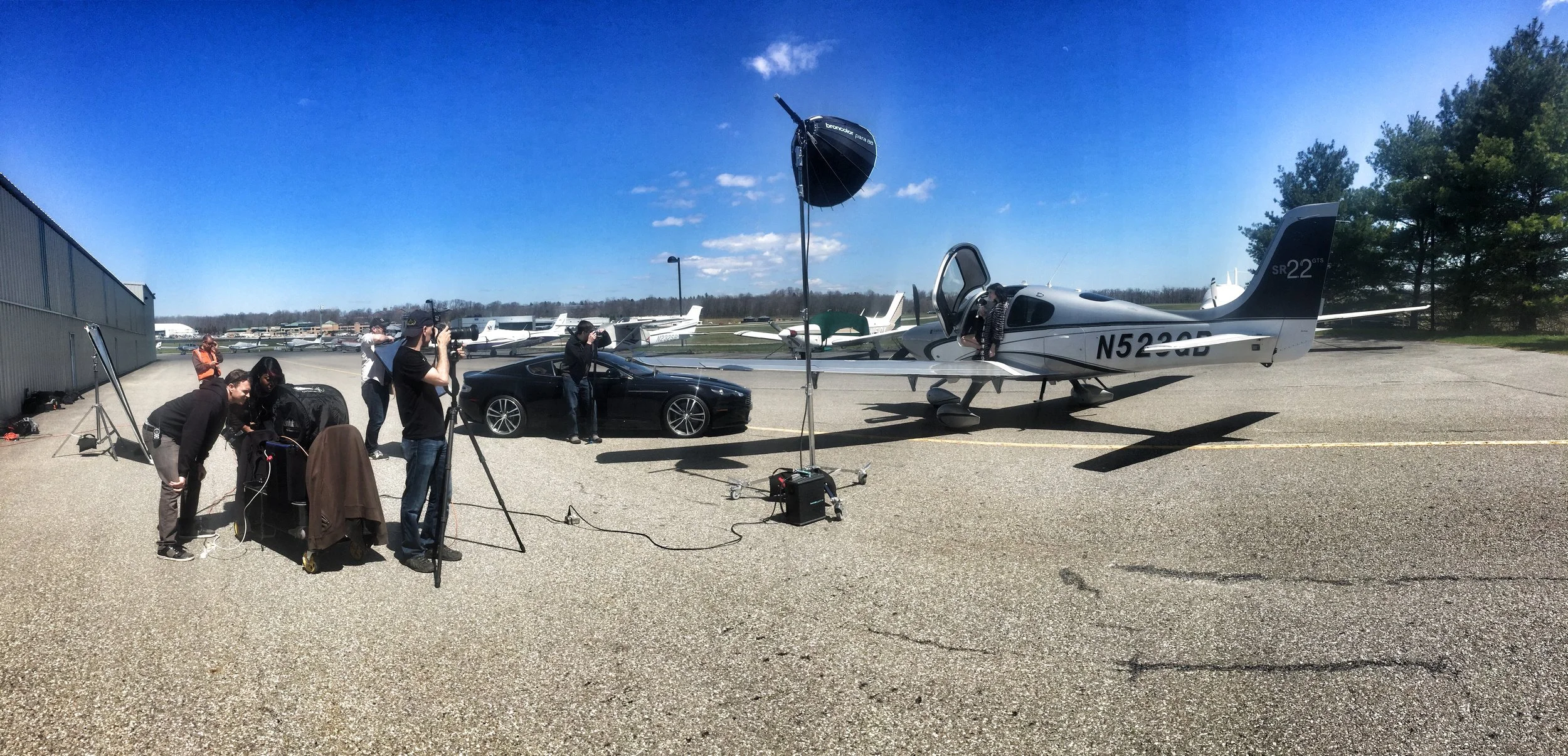 A photoshoot taking place at an airport with a small airplane, a black car, and a group of people setting up cameras and equipment.