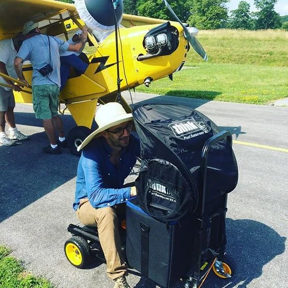 Man sitting on a golf cart with a large sun hat, while several people work on a small yellow airplane on a grassy airstrip.
