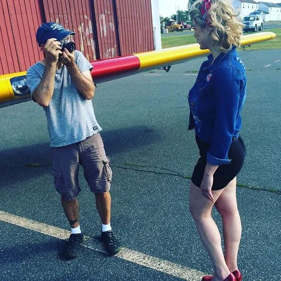 A woman posing for a photo taken by a man holding a camera. The woman is wearing a denim jacket and high heels, and has blonde hair with a colorful headband. The man is wearing a gray t-shirt, cargo shorts, and a cap, standing in front of a red and y