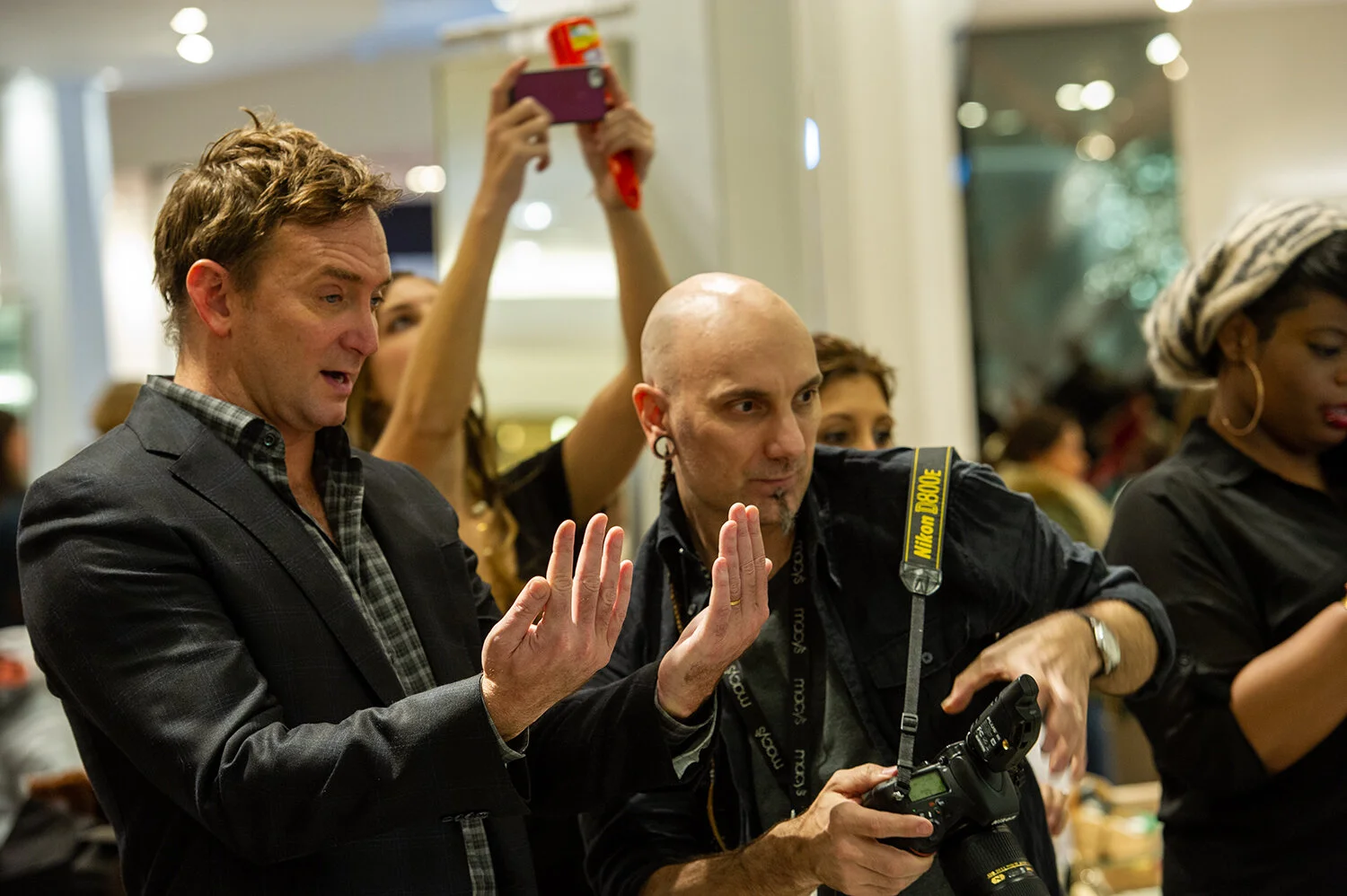 Two men at a social event, one in a dark blazer and the other with a Nikon camera, both gesturing with their hands, with a woman taking a photo in the background and other people in the crowded indoor space.