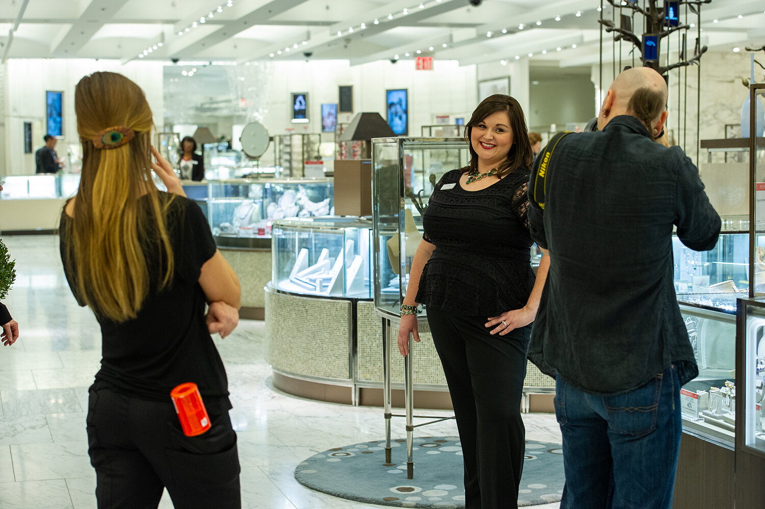 A woman with dark hair, wearing a black top and pants, standing inside a jewelry store, smiling at a customer or a photographer.