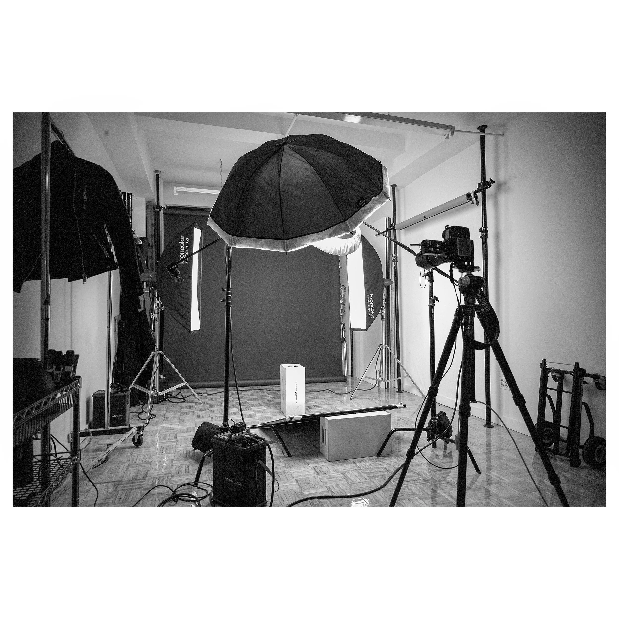 Photography studio setup with umbrellas, softboxes, camera on tripod, backdrop, and lighting equipment on a wooden floor.