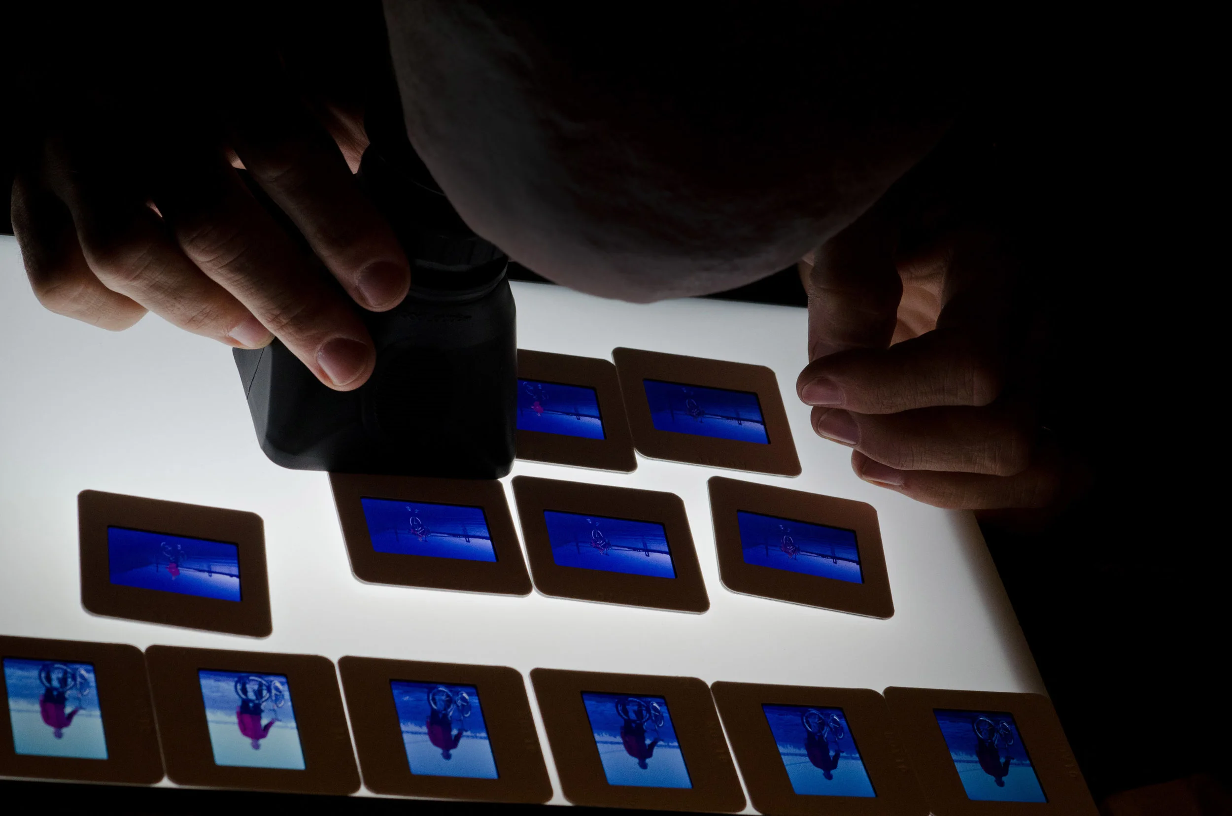 Person flipping through photographic slides on a lightbox, with a camera in hand, creating a stereoscopic or 3D effect.