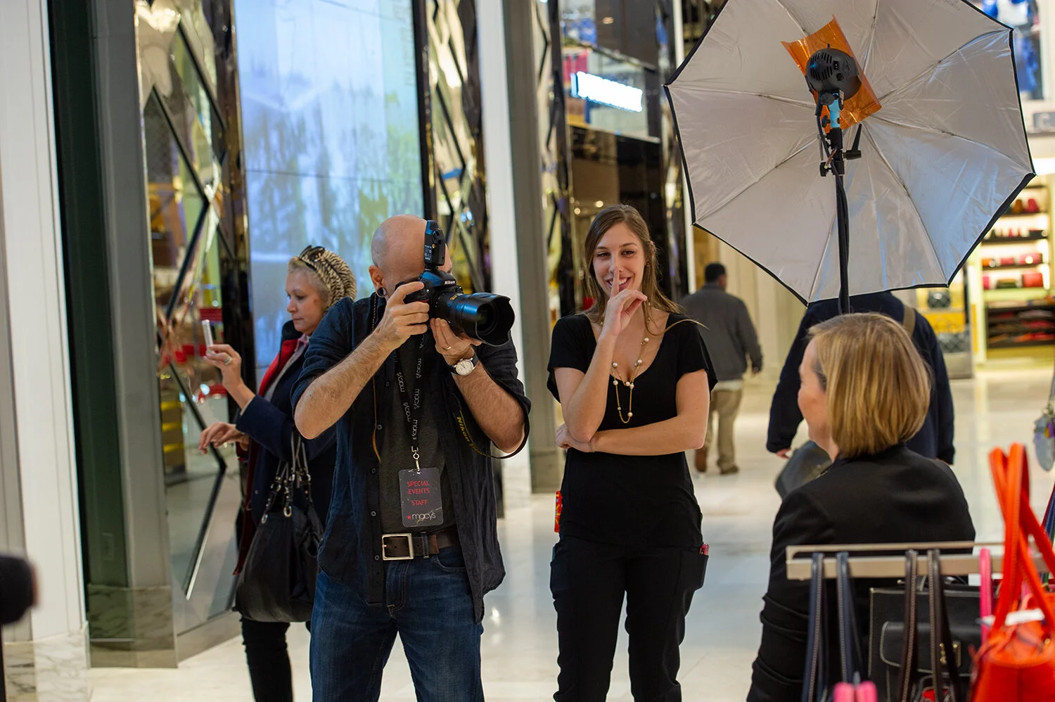 Photographer taking pictures of smiling woman in shopping mall, with a woman in the background looking at her phone, and a woman seated at a table with a large umbrella overhead.
