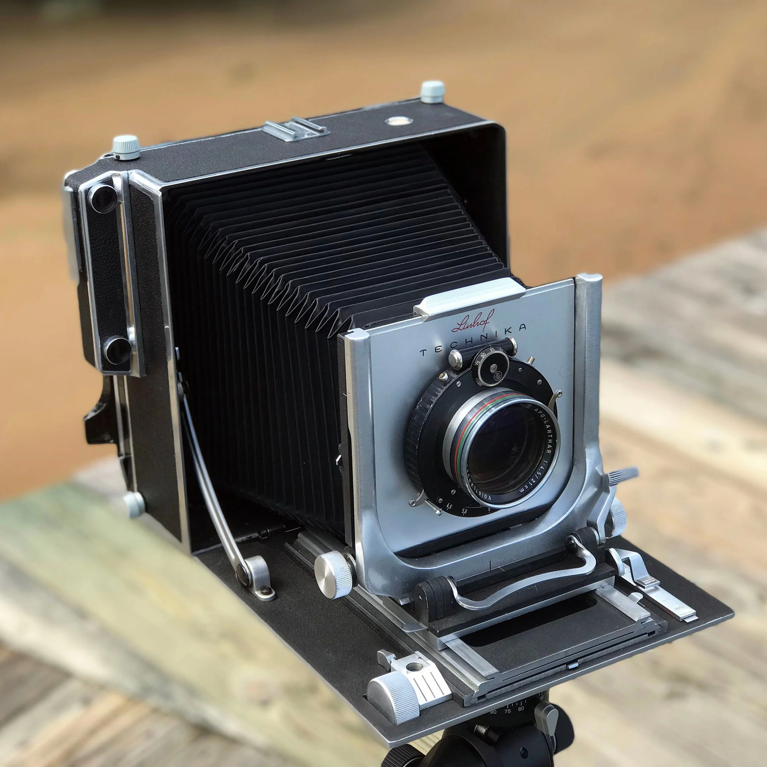 A vintage Linhof Technika large format camera with a bellows and a lens, mounted on a tripod on a wooden surface.