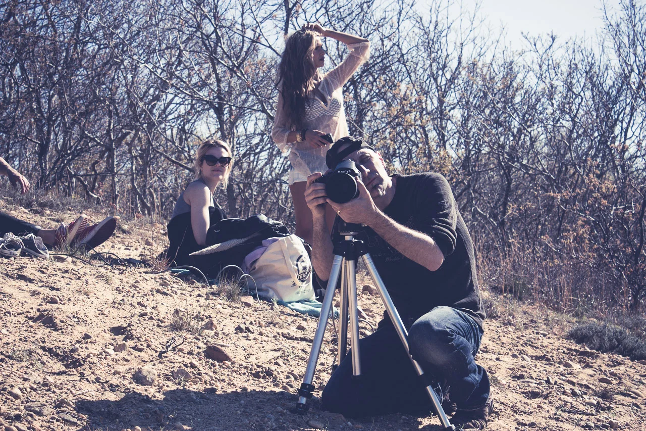 A man with a camera on a tripod taking a photo. Two women sitting and standing on a rocky hill with backpacks and shoes nearby, under leafless trees.