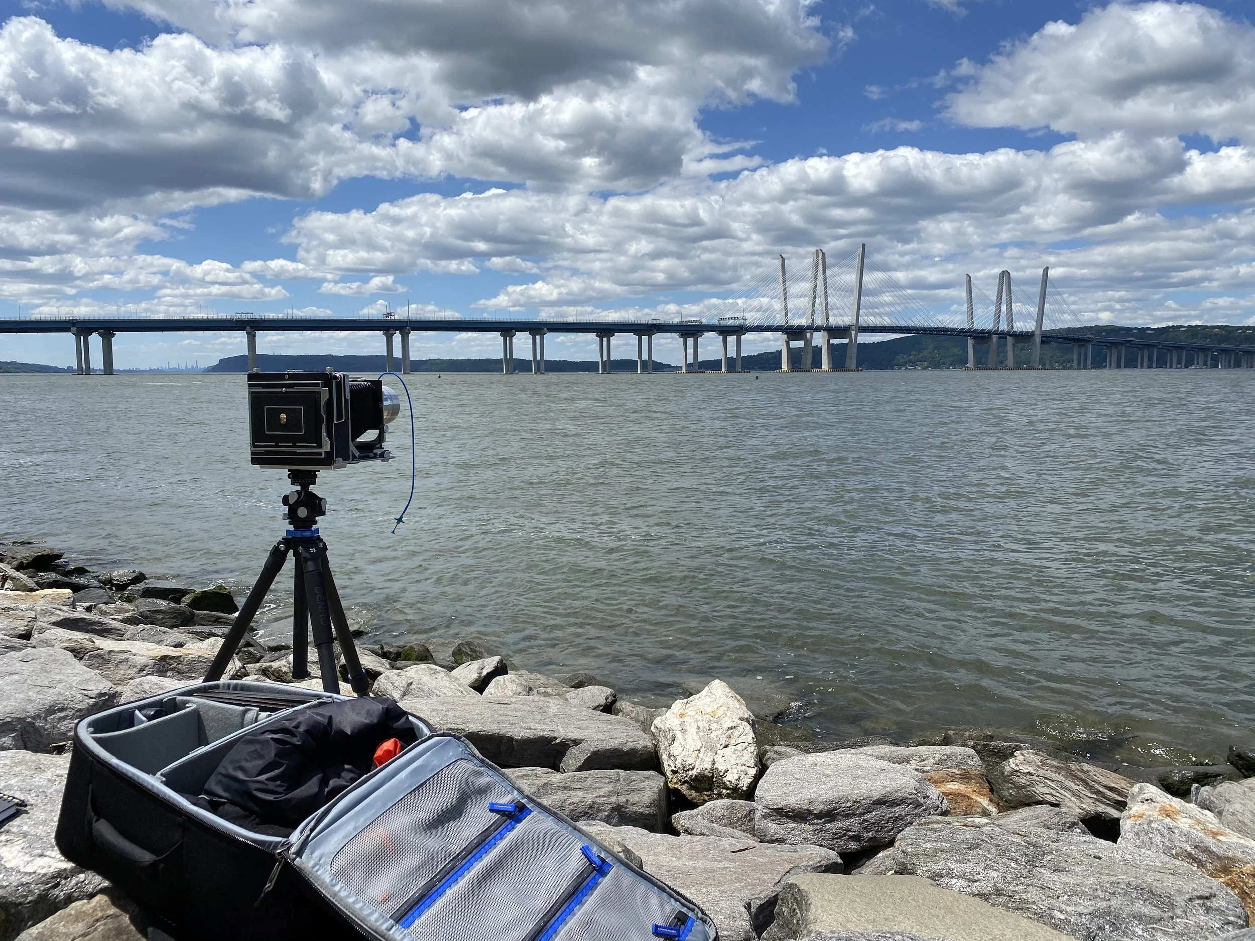 A camera on a tripod set up on rocky shoreline by the water with a bridge and partly cloudy sky in the background.