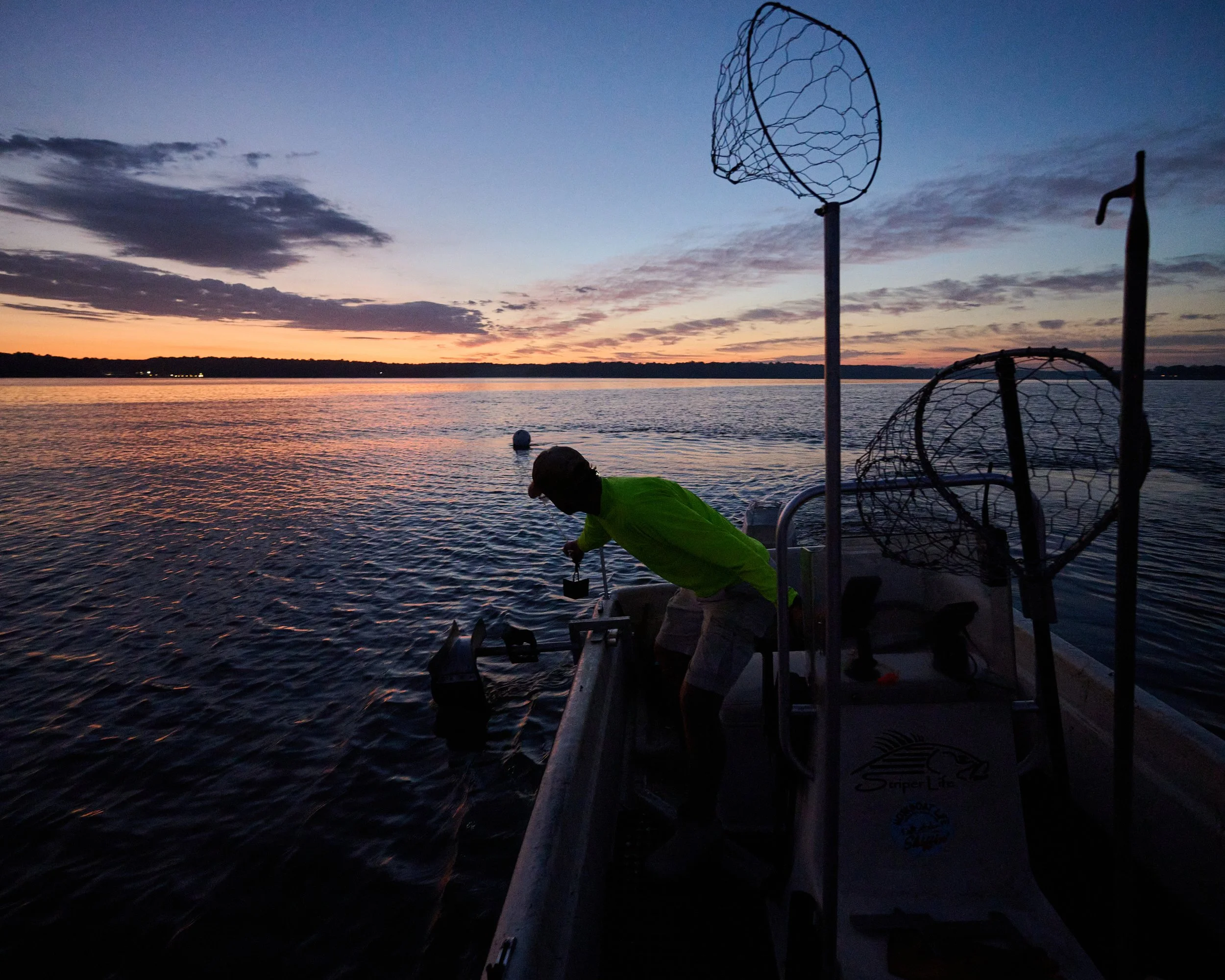 A person on a boat fishing at sunset, with the sky painted in shades of orange, pink, and purple, and the calm water reflecting the colors.