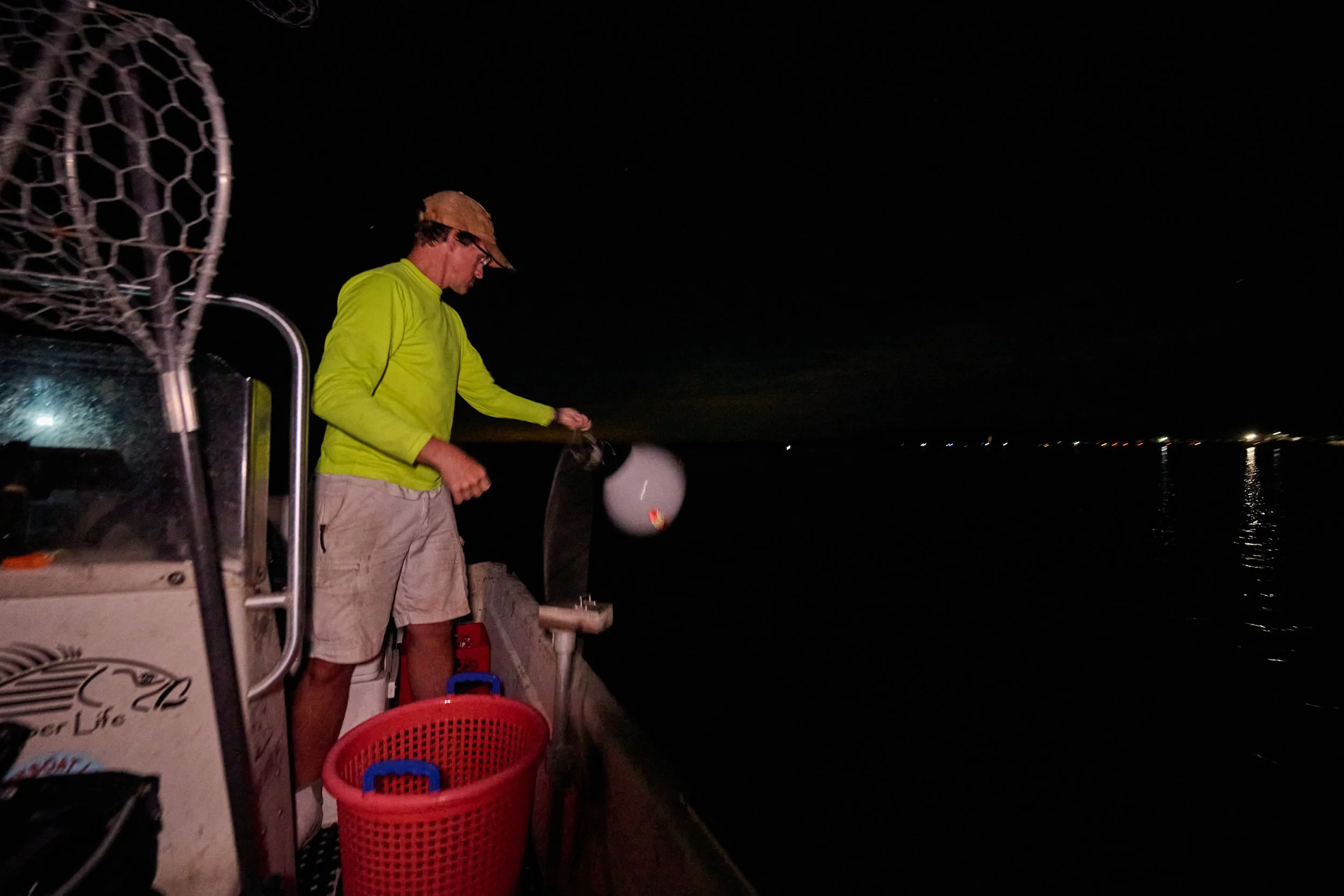 A man in a bright yellow shirt and beige shorts on a boat at night, holding a fishing net over the water with city lights visible in the distance.