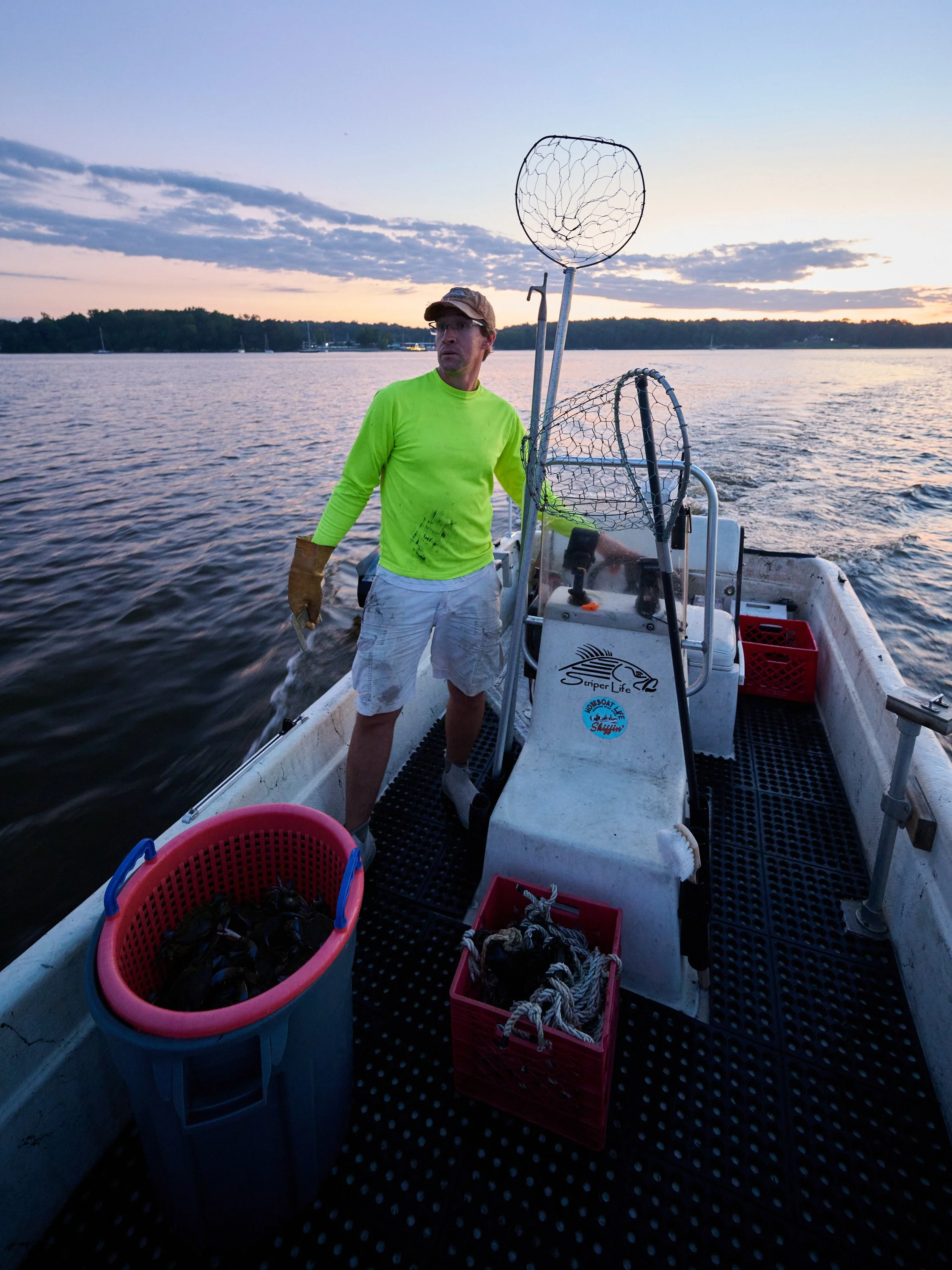 A man in a neon green long-sleeve shirt, white shorts, and a baseball cap on a boat at sunset, holding fishing gear with baskets of caught fish nearby, on a body of water with trees in the background.