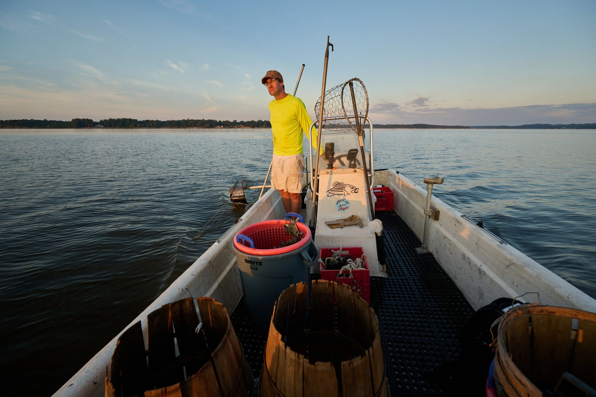 A man in a yellow shirt and shorts standing on a small boat on a calm lake at sunset, surrounded by buckets and fishing gear.