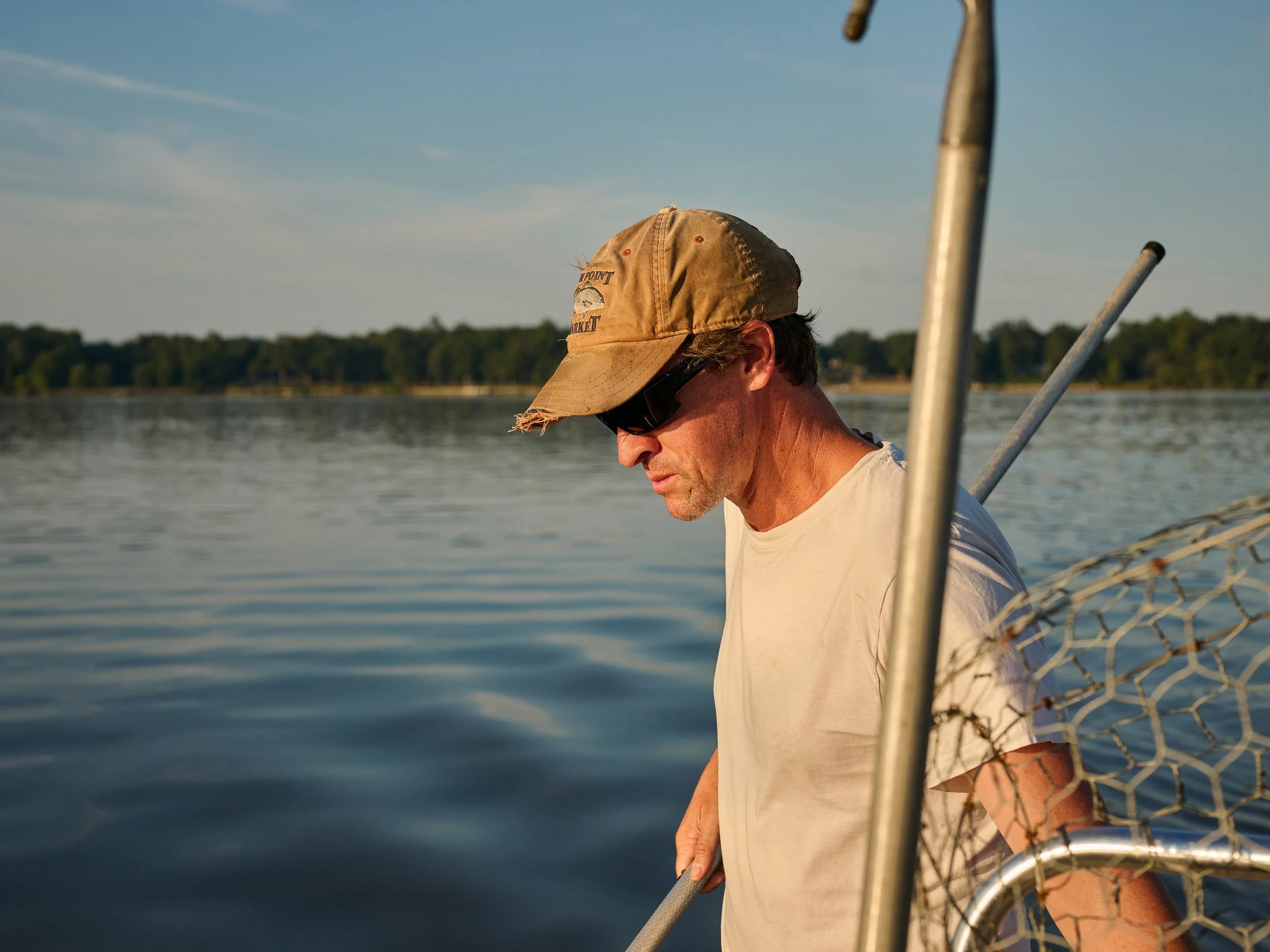 A man with a tan cap, sunglasses, and a white t-shirt is fishing on a lake during sunset. He is looking down and holding a fishing net.
