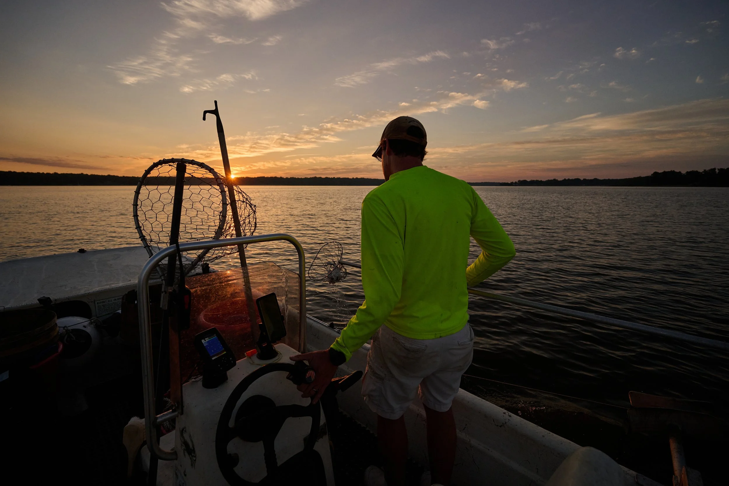A man in a neon yellow shirt and gray shorts standing on a boat at sunset, holding a fishing rod while looking at the water.
