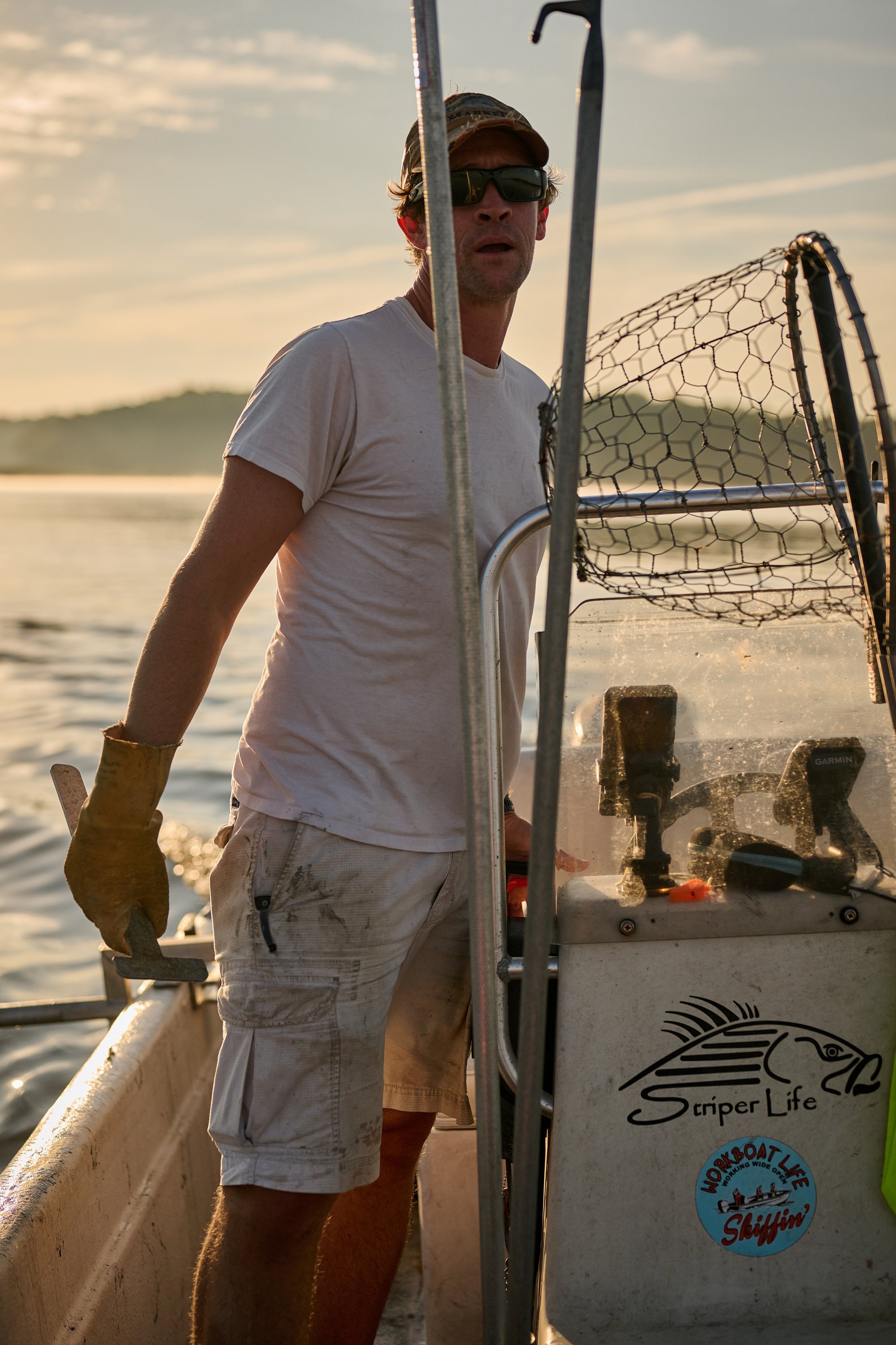 A man standing on a boat, wearing a white t-shirt, beige cargo shorts, yellow gloves, a cap, and sunglasses, with boats and water in the background during sunset.