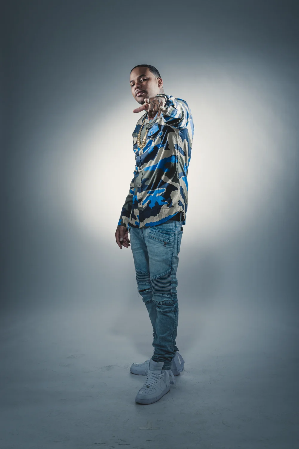 A man in a camouflage jacket, ripped jeans, and white sneakers posing with a confident expression and pointing towards the camera in a photo studio.