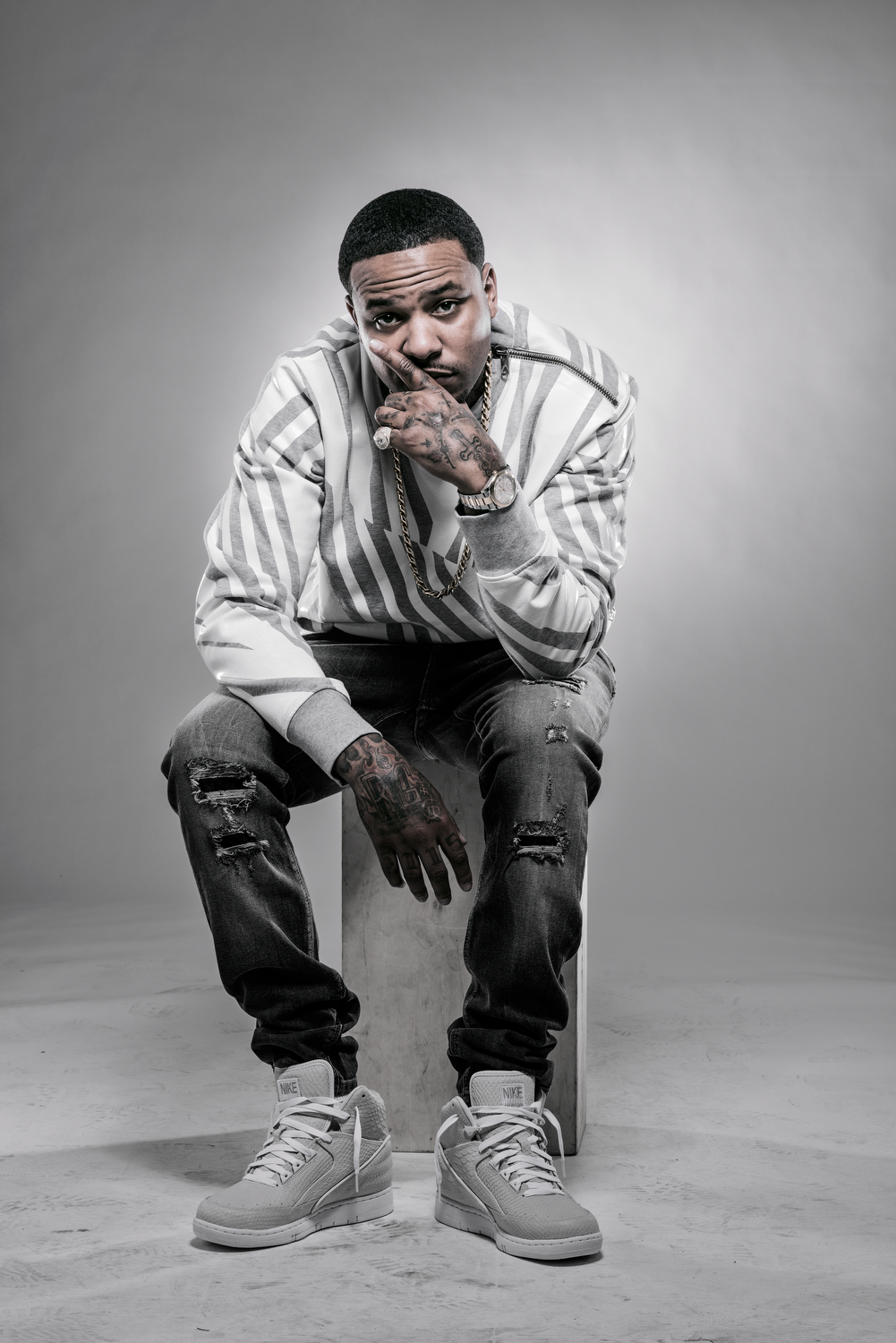 Black and white photo of a man sitting on a box, wearing a striped hoodie, ripped jeans, and Nike sneakers, with tattoos, jewelry, and a watch, striking a pose with one hand near his face.