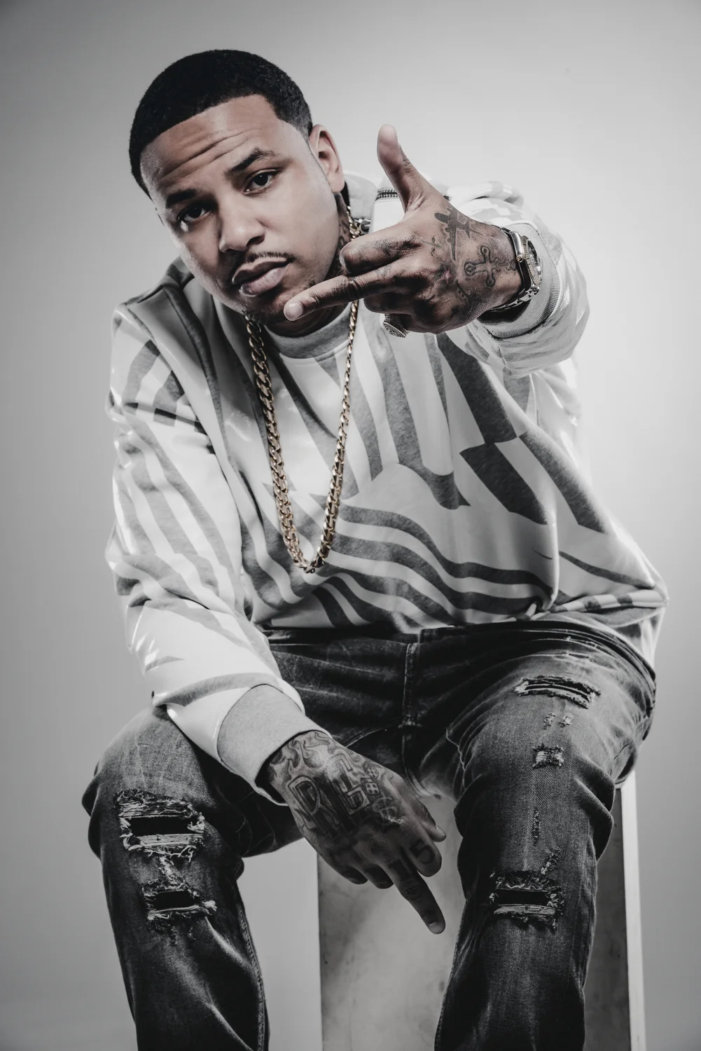 A man with short hair and tattoos on his hands is sitting and making a gesture with his right hand, pointing his index finger and thumb in a gun shape. He wears a patterned hoodie, ripped jeans, a gold chain, and a watch.