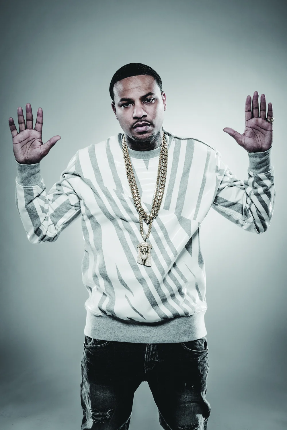 A young man with short hair and a serious expression wears a white and gray striped sweatshirt and multiple gold chains, holding both hands up with palms facing out against a plain background.