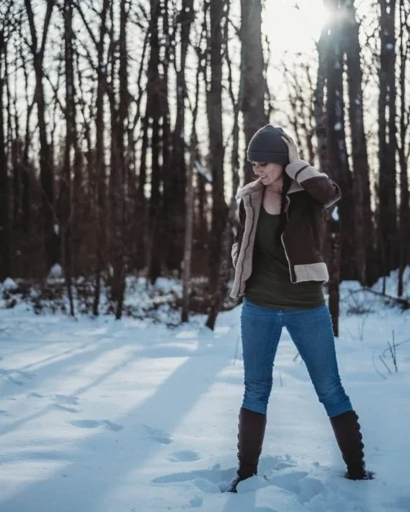 A woman standing in a snowy forest, wearing a jacket, beanie, and jeans, with the sun shining through the trees in the background.