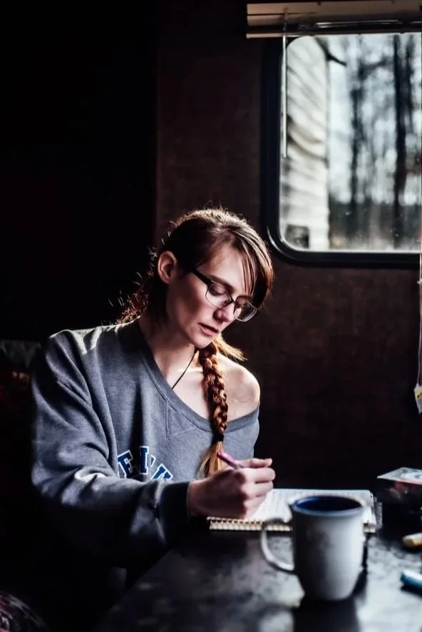 Young woman with glasses and braided hair writing in a notebook at a table near a window showing trees outside.