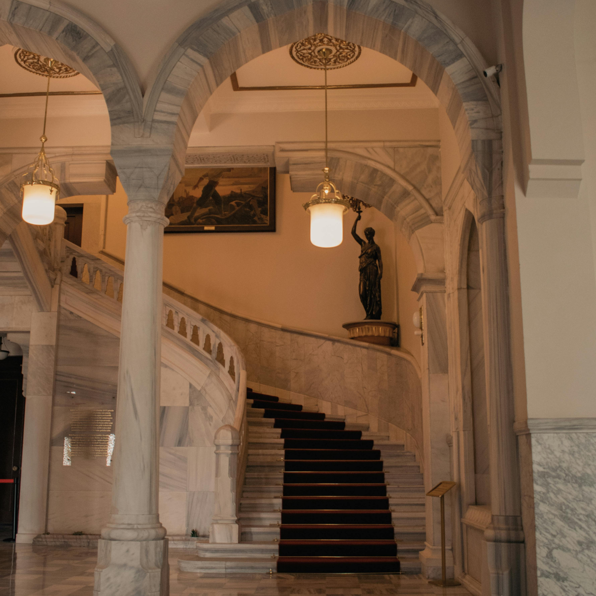Marble staircase with a black runner, ornate arches, a bronze statue of a woman holding a torch, and warm lighting fixtures in an elegant building interior.