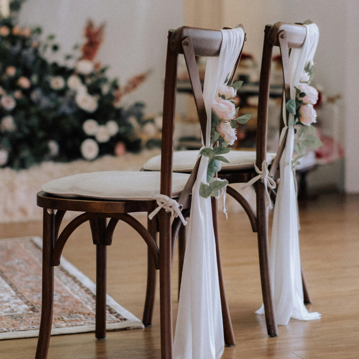 Two wooden chairs decorated with white fabric and pink roses for a wedding or special event, positioned on a hardwood floor with a floral arrangement in the background.