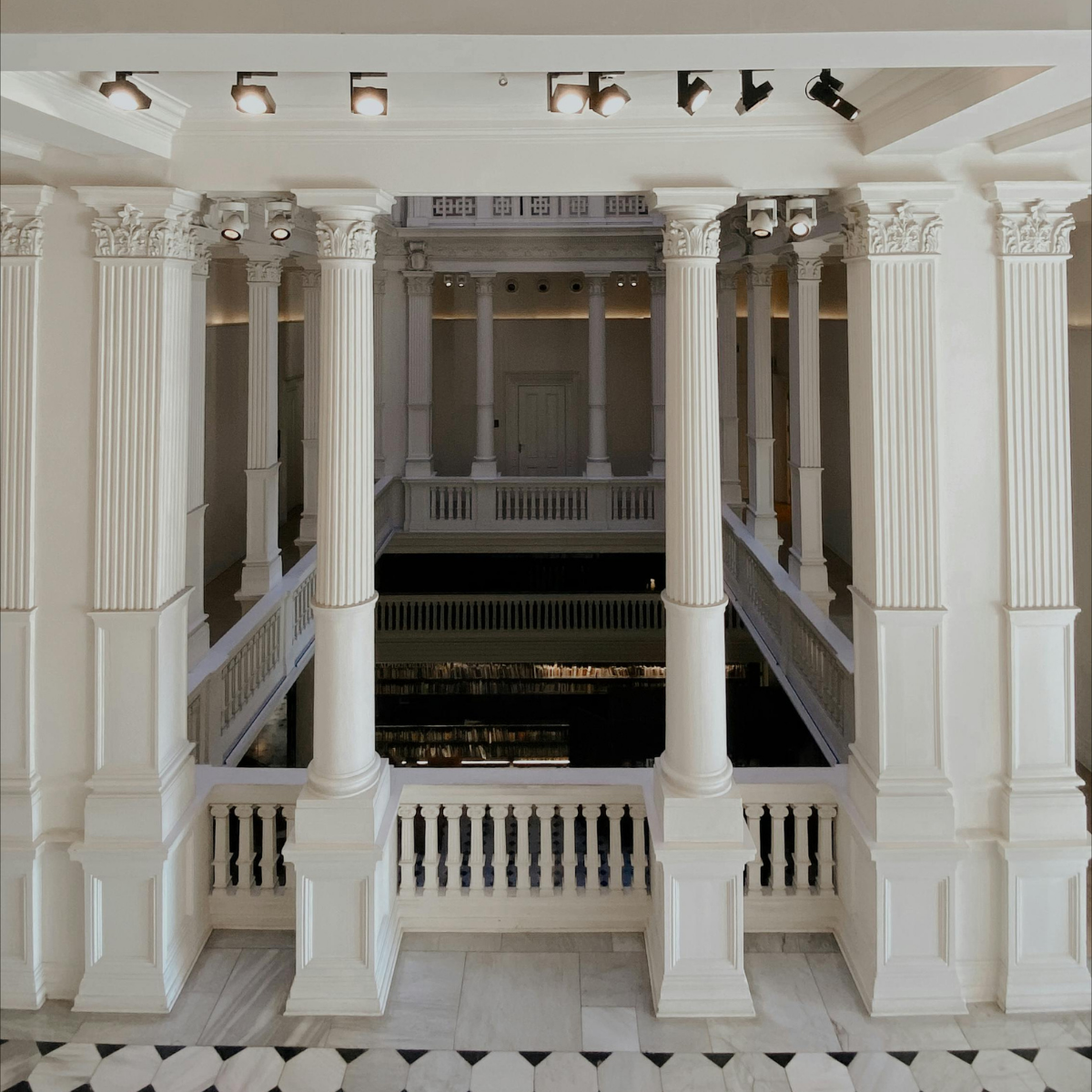 Interior of a building with white columns, detailed capitals, and a balcony railing, featuring a spacious, classical architectural style.