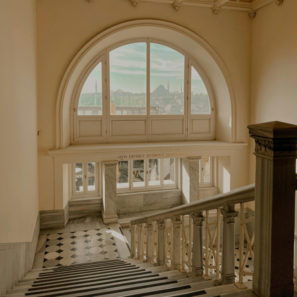 View from the top of a staircase looking out a large arched window with a cityscape in the distance, including minarets and domes.