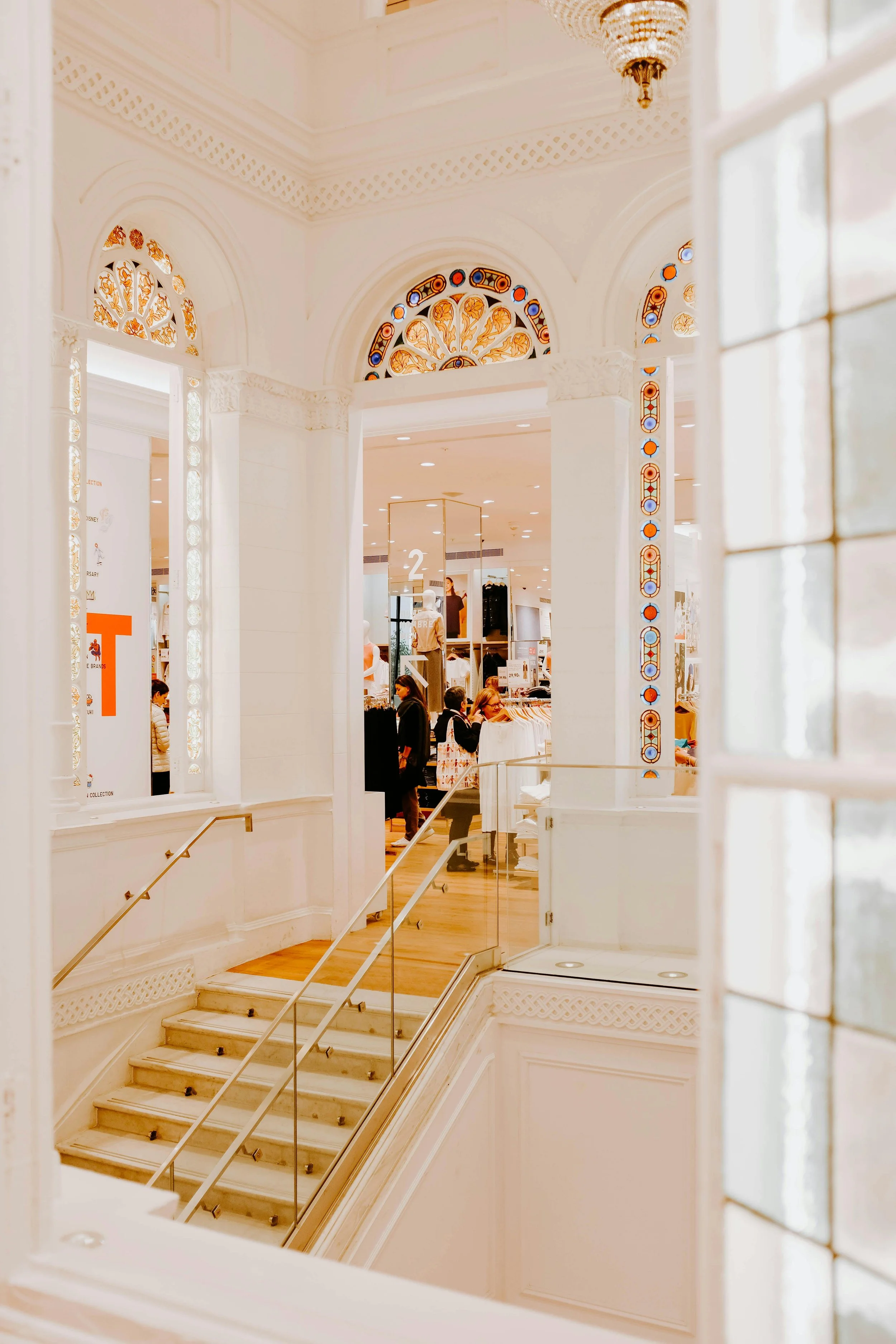 View of a shopping mall interior with ornate white walls, stained glass decorative arches, and a staircase leading down. People are shopping and browsing in the background.
