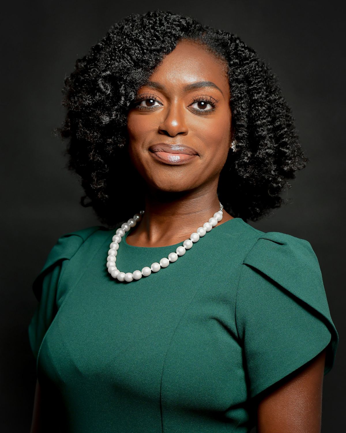 Myriam Santos is wearing a green dress, a pearl necklace, and pearl earrings, posing against a black background.