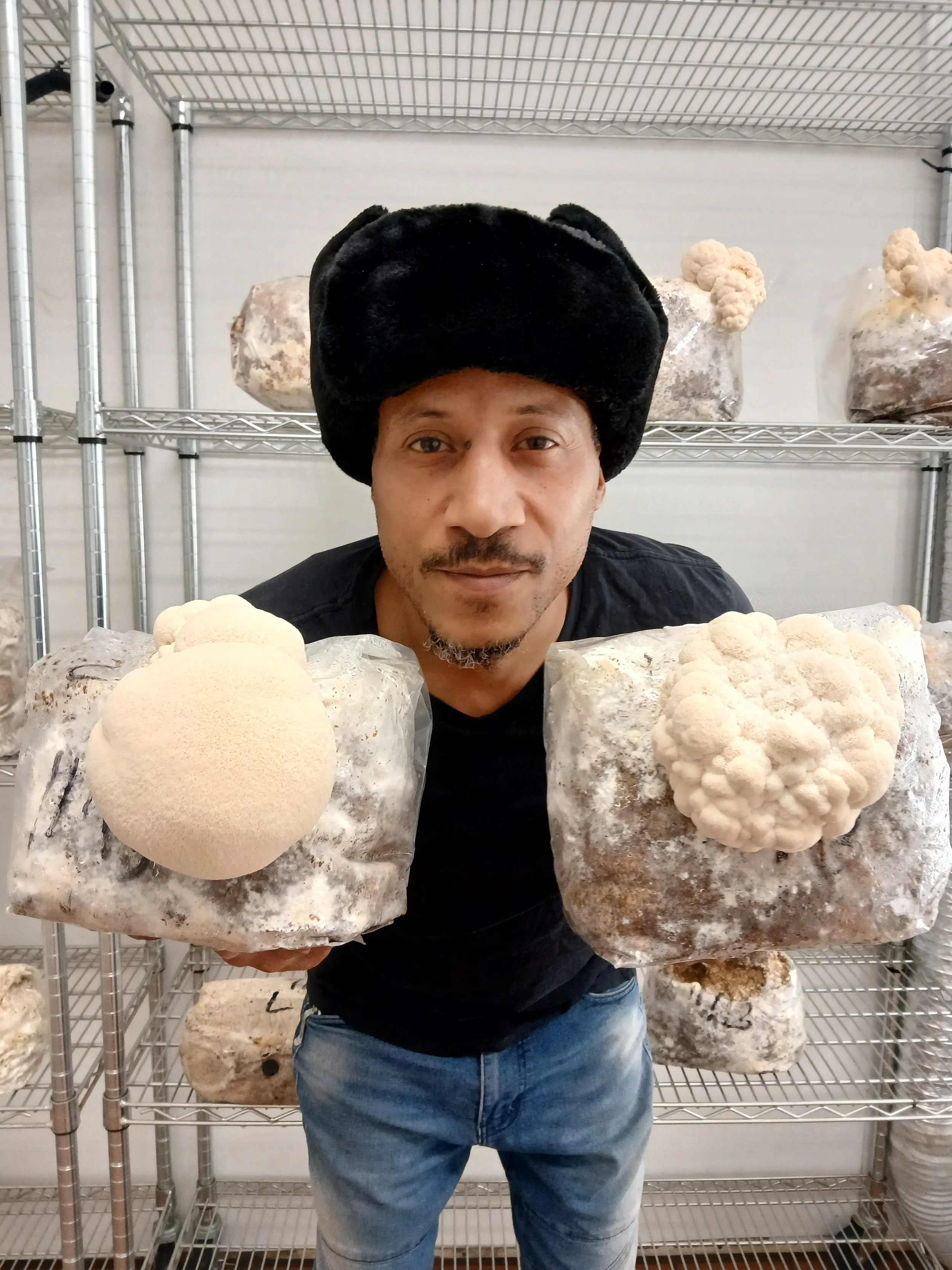 A man wearing a black hat holding two large blocks of mycelium with mushroom growth, standing in front of metal shelving with more blocks of mushroom cultivation substrate.