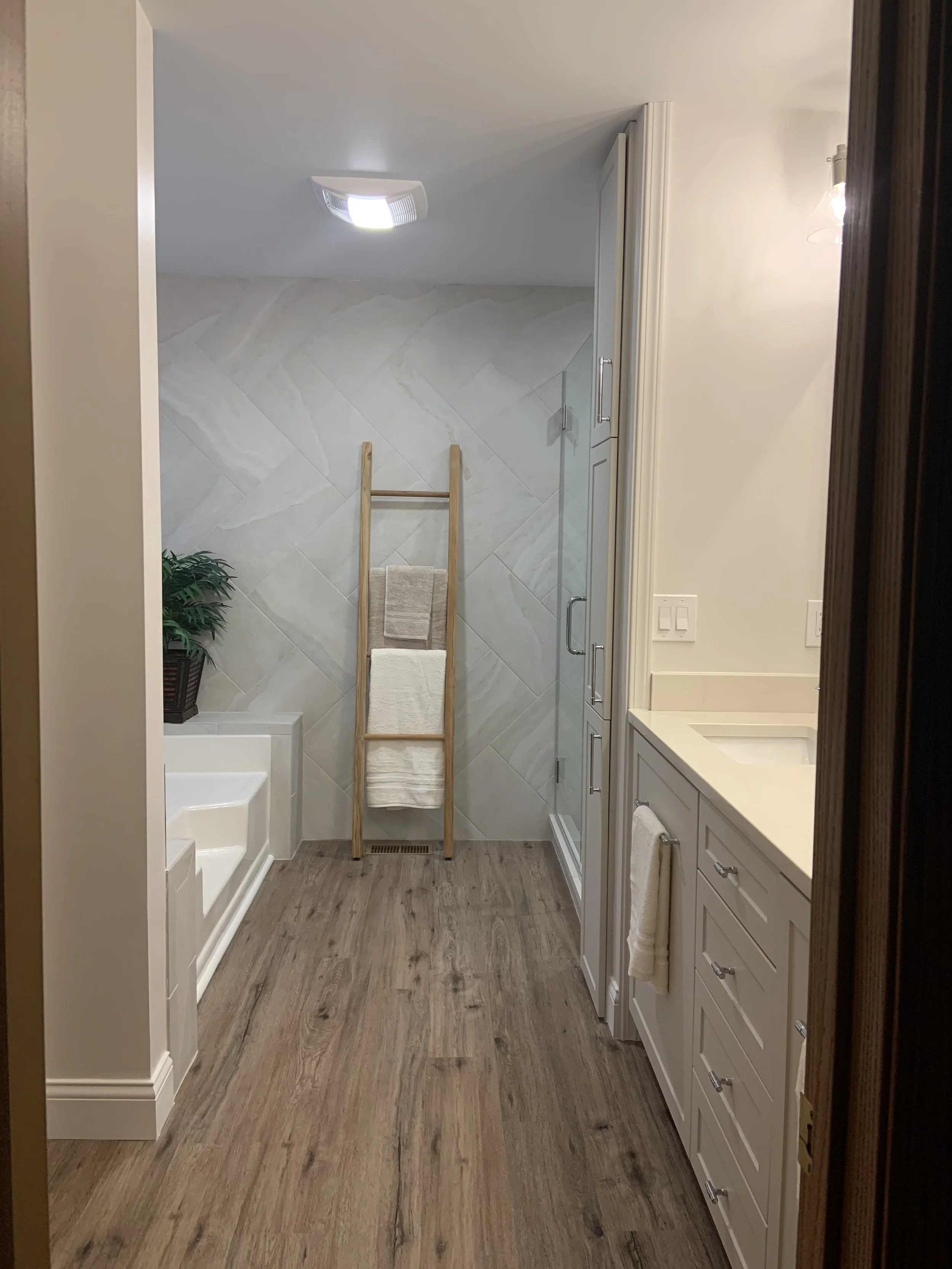 Bathroom with a bathtub on the left, wooden towel rack with towels in the center, and a shower with glass door on the right. White cabinets and a sink on the far right, wooden floor, and a potted plant on the left.